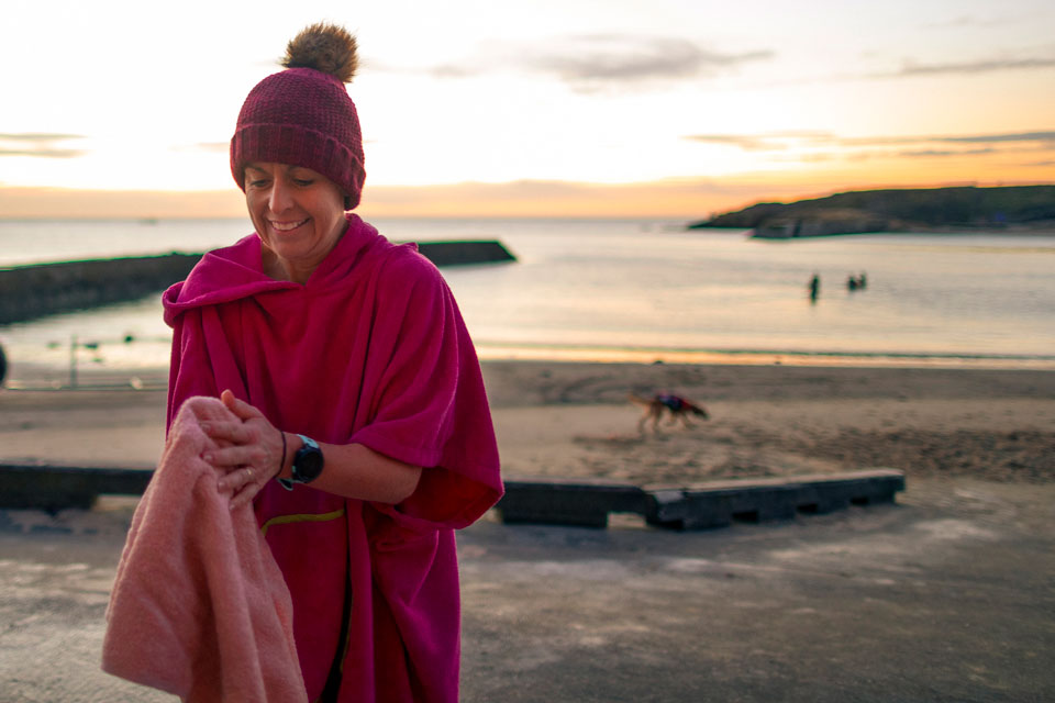 Open water swimmer standing on the beach wearing warm layers and a hat, holding a mug, after a swim in the sea
