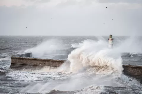 Storm waves crash over harbour wall and lighthouse in stormy seas