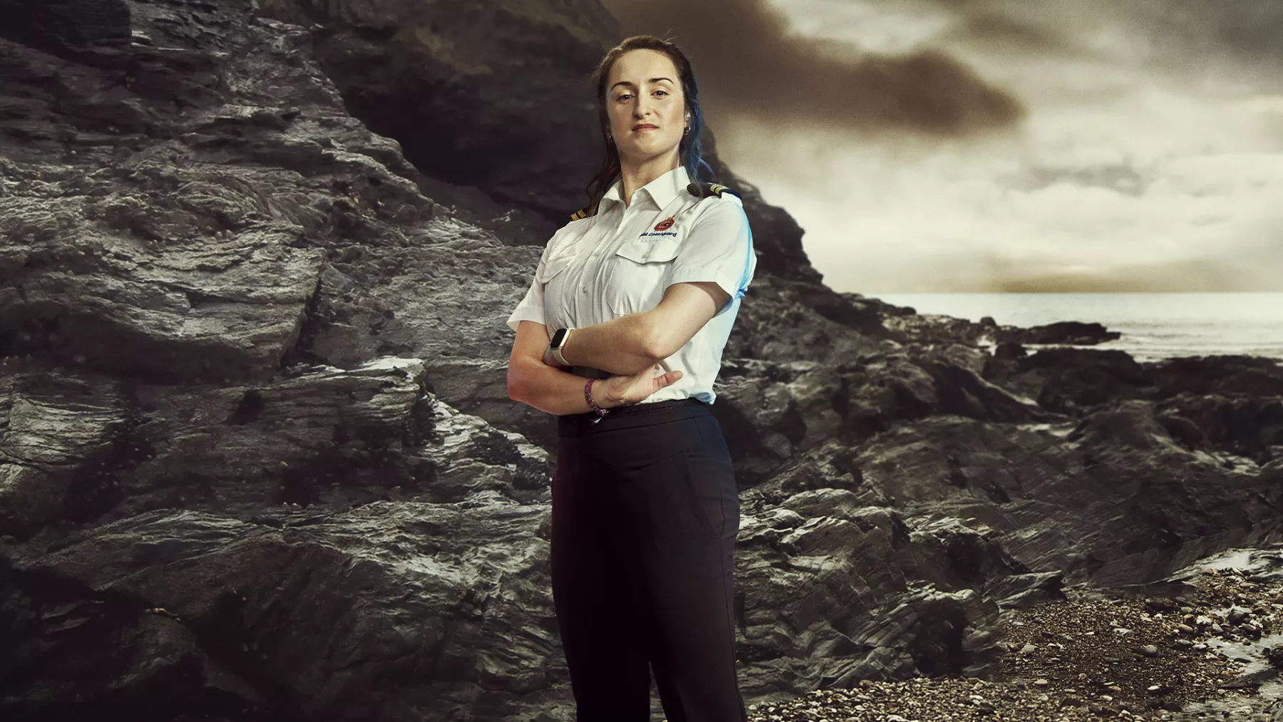 A female Coastguard officer stands with her ams folded on a grey, rocky landscape. She is wearing an official white shirt with the HM Coastguard logo and epaulettes.