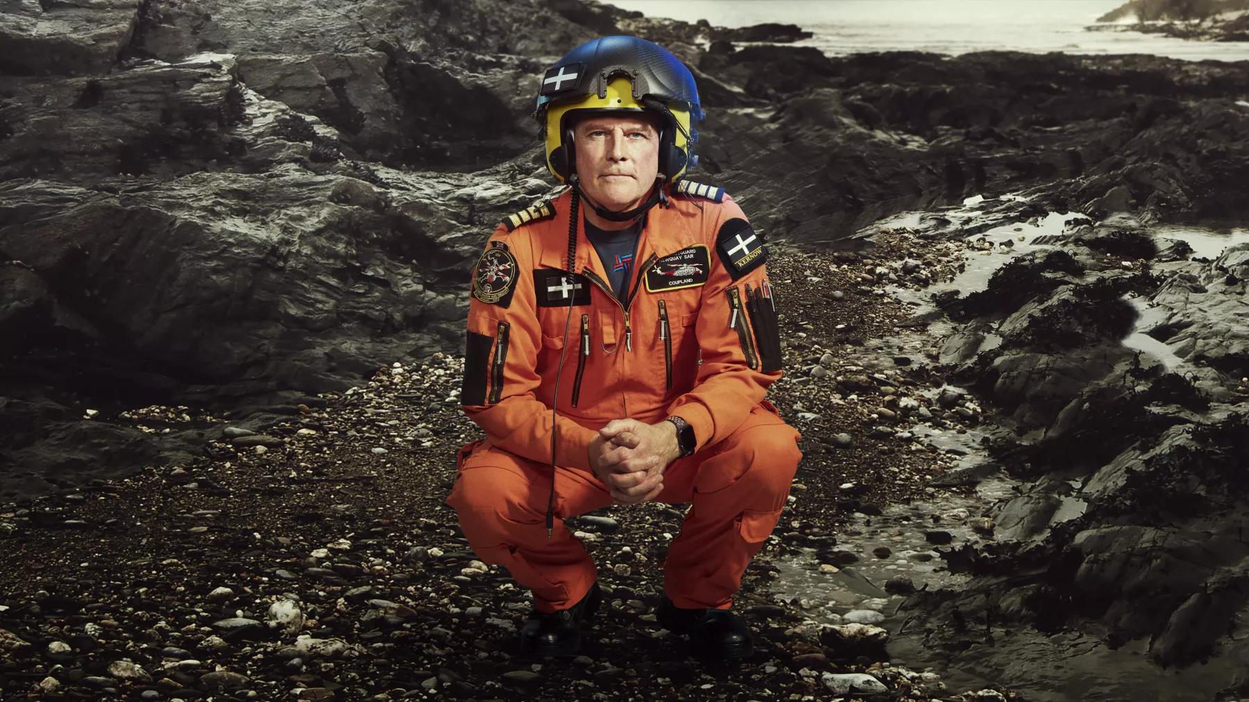 Crouched down helicopter crew member on rocky terrain in orange coveralls and helmet