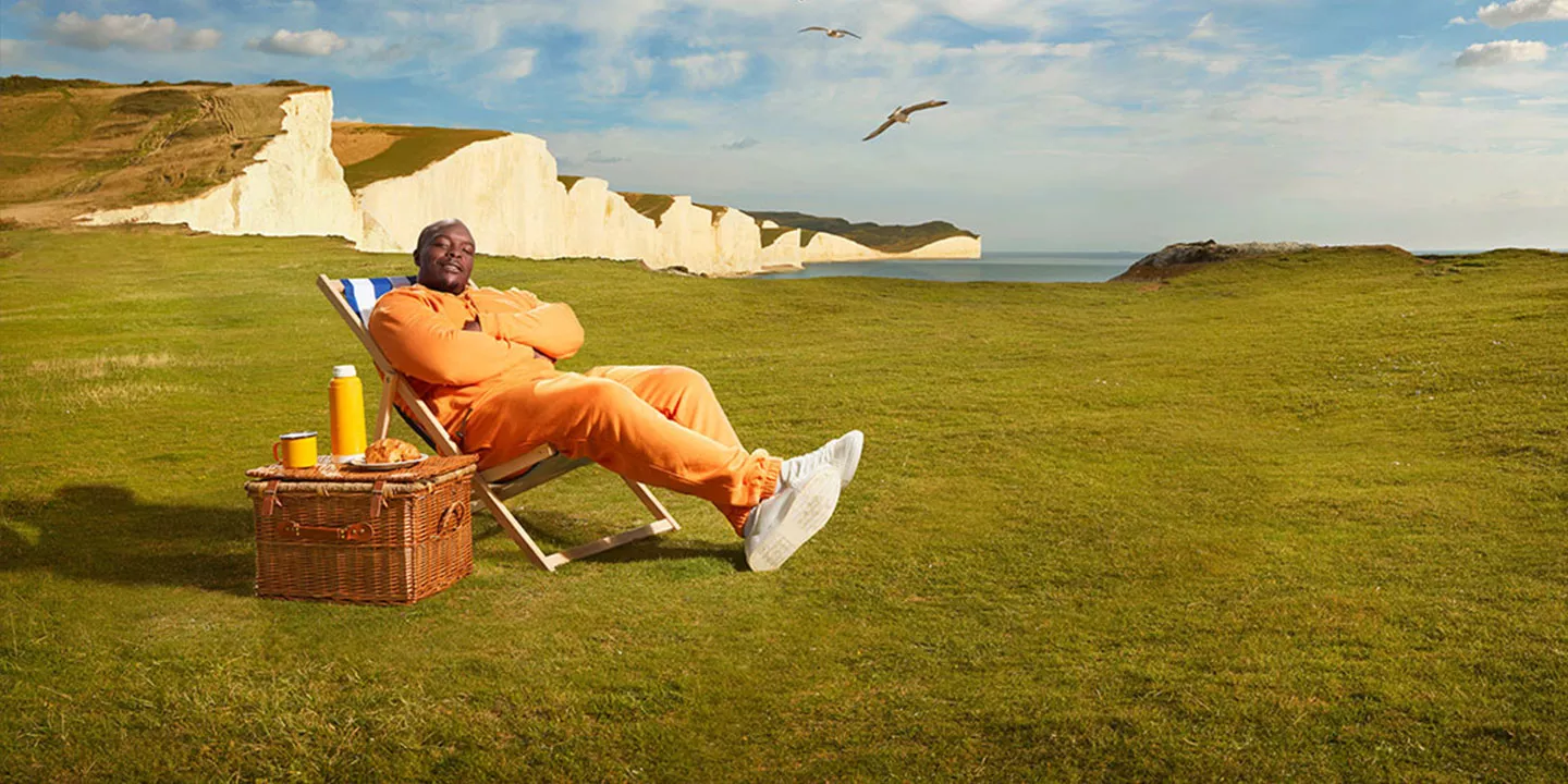 A man dressed in a bright orange jumpsuit relaxes on a deck chair with the rocky coast in the background