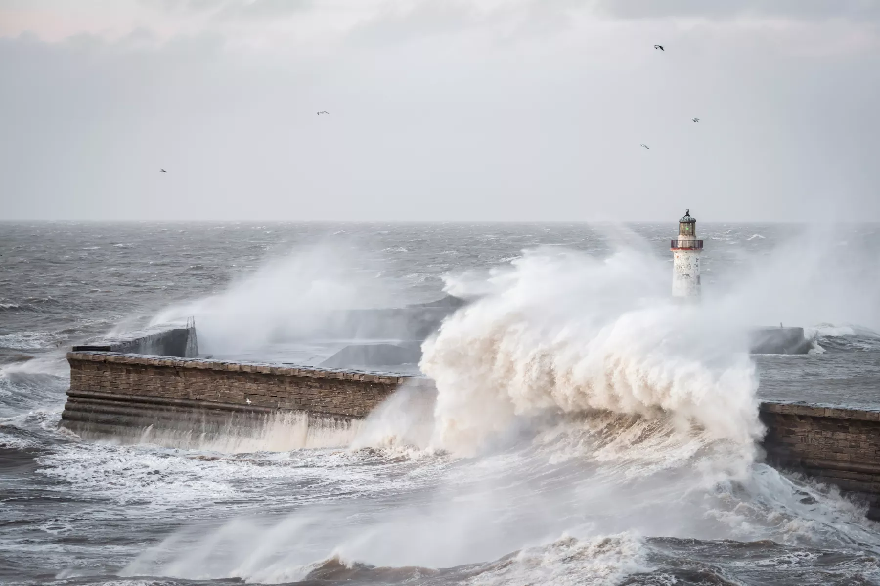 Storm waves crash over harbour wall and lighthouse in stormy seas
