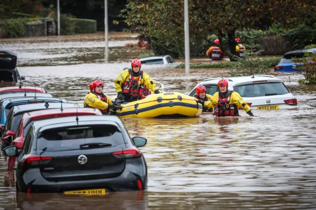 Coastguard flood rescue officers in flooded street with inflatable used to rescue people, floating past parked cars part-submerged
